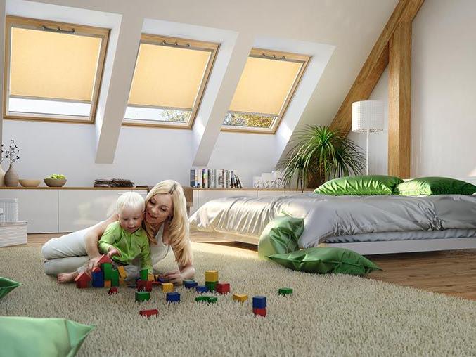Egy anya játszik gyermekével a hálószoba padlóján, a háttérben 3 fa tetőablak, szinte zárt redőnyökkel.
